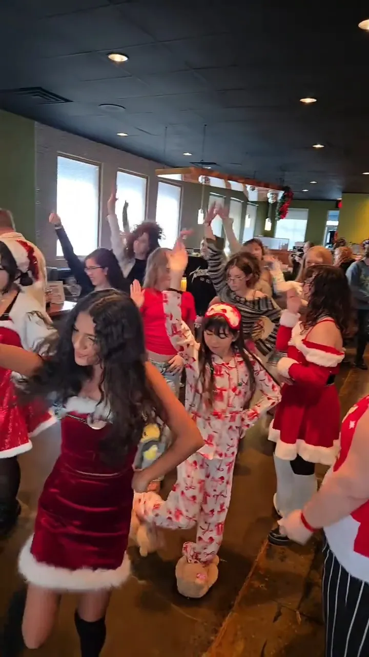 A crowd of kids and adults dance with arms raised inside a decorated room, several wearing red Santa dresses and holiday pajamas, at Knox Pop Con.