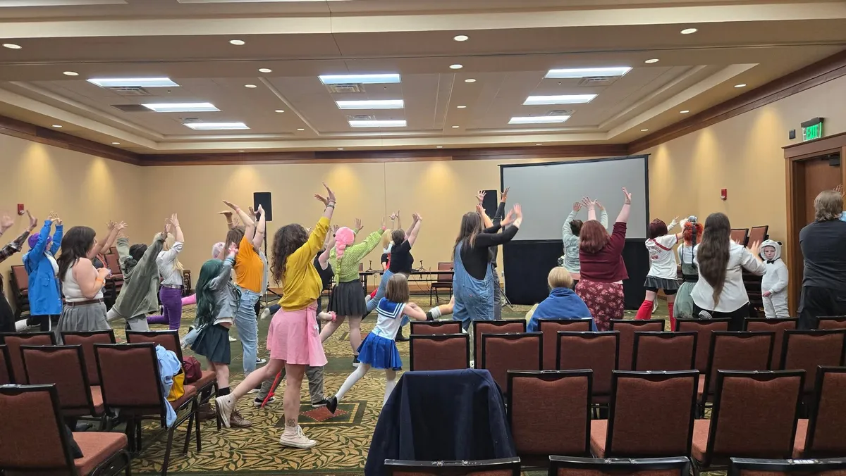 A large group in cosplay and casual outfits raises their arms in unison during a dance workshop inside a carpeted ballroom with a projector screen and rows of stacked chairs, at Knox Pop Con.