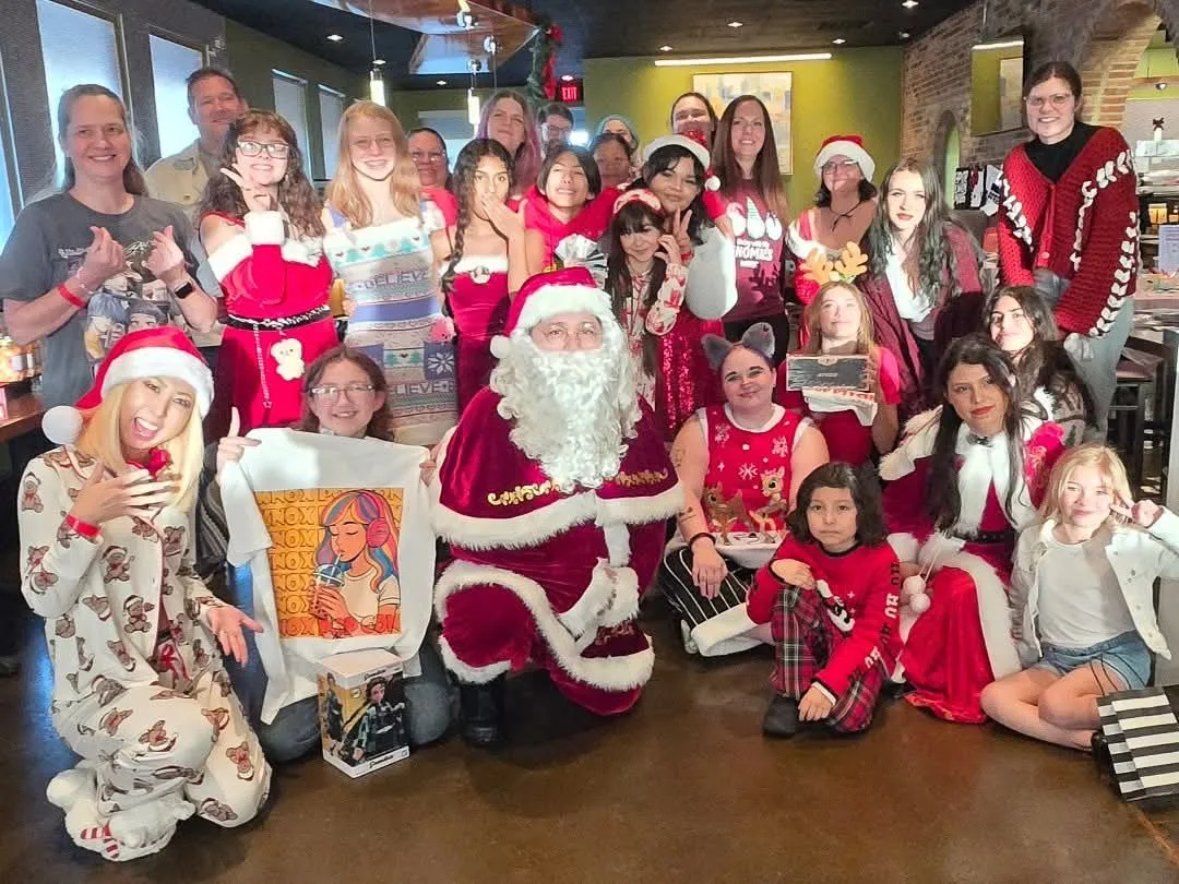 A large group in Santa outfits, festive sweaters and cosplay poses together on the floor of a brick-walled restaurant, with one person holding up a Knox Pop Con art tee, at Knox Pop Con.